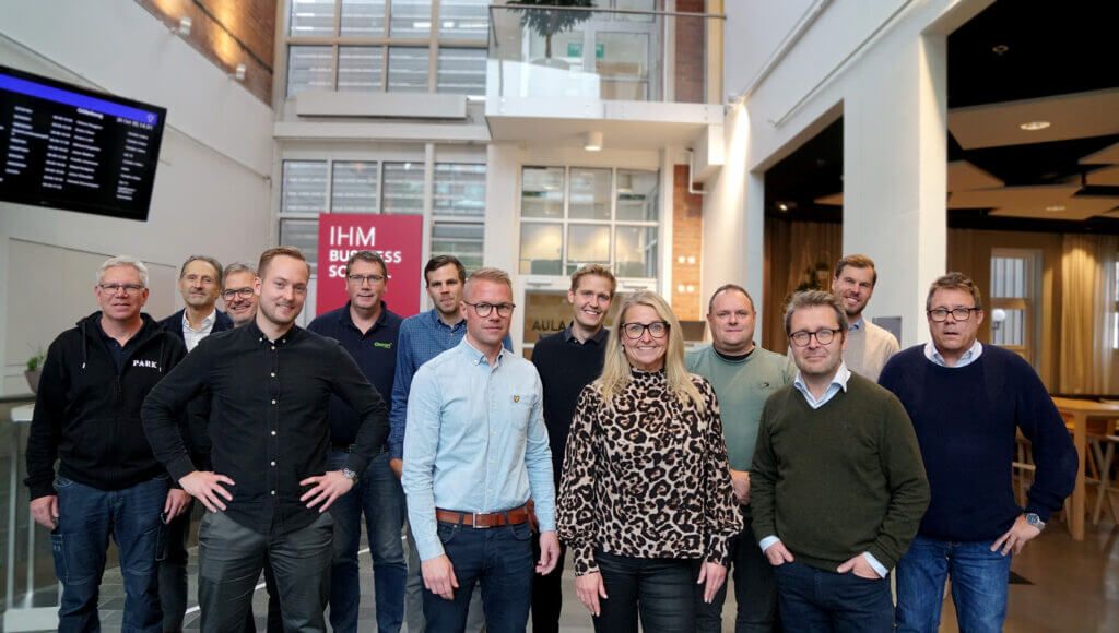 Grupp på tretton personer som står i ett kontorsutrymme, några bär informella klädsel och andra i business casual, ler mot kameran.